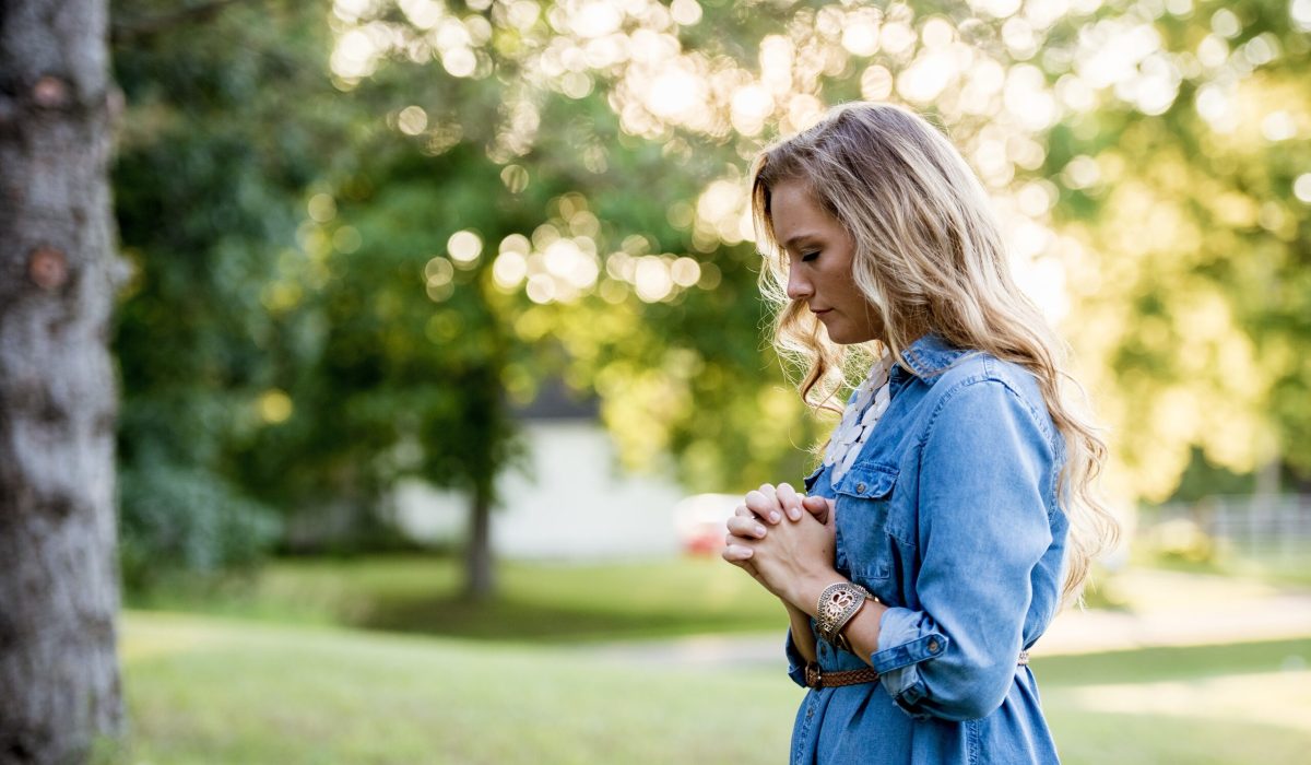 A woman wearing a blue dress and praying in a garden under sunlight with a blurry background